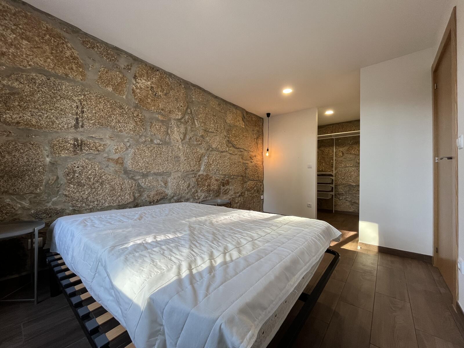 Quarto do T1 Casa Linho com a parede de pedra de granito mantida à vista e soalho em madeira.